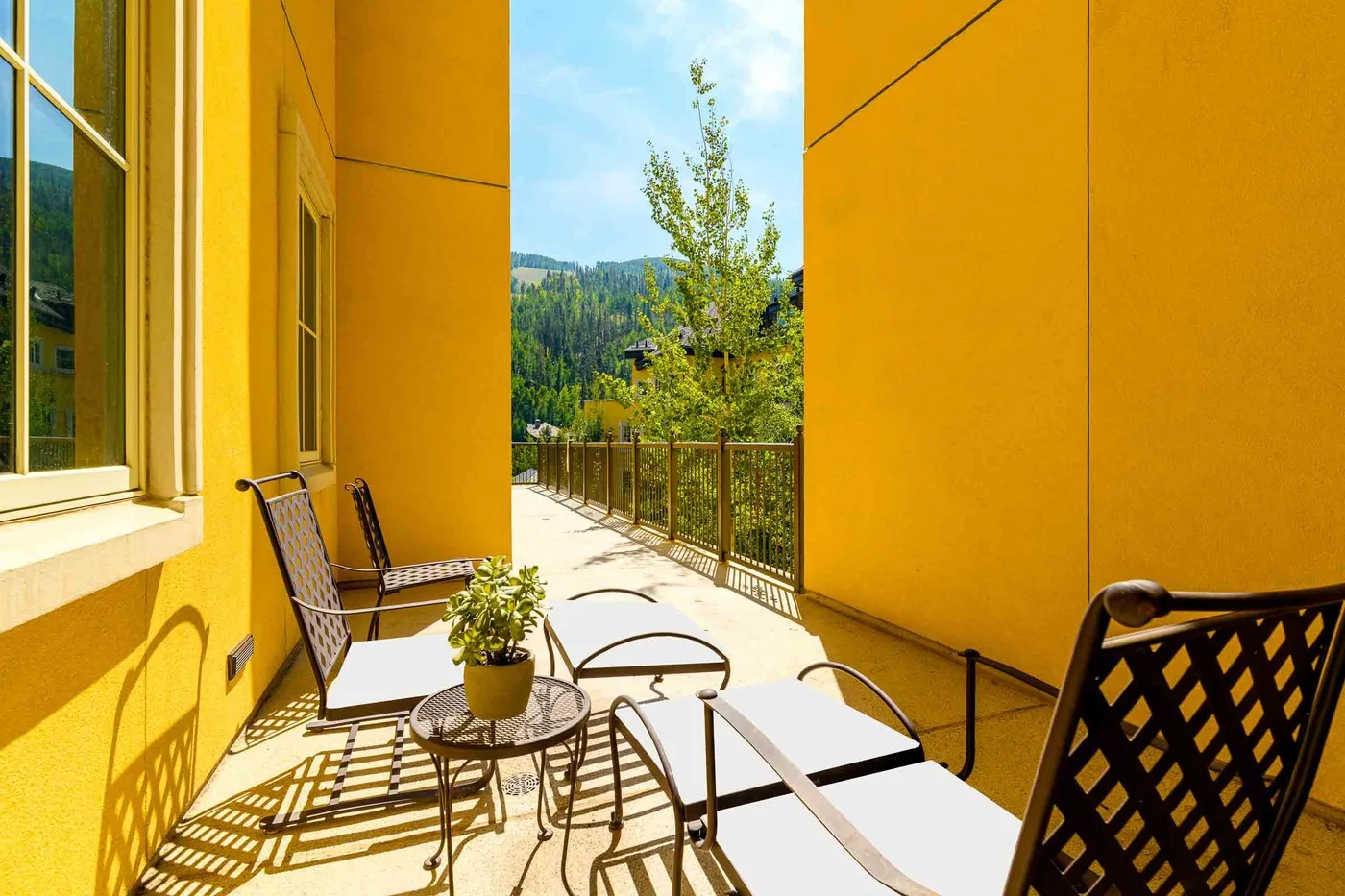 A sunny outdoor patio with seating and a view of trees beyond the railing.