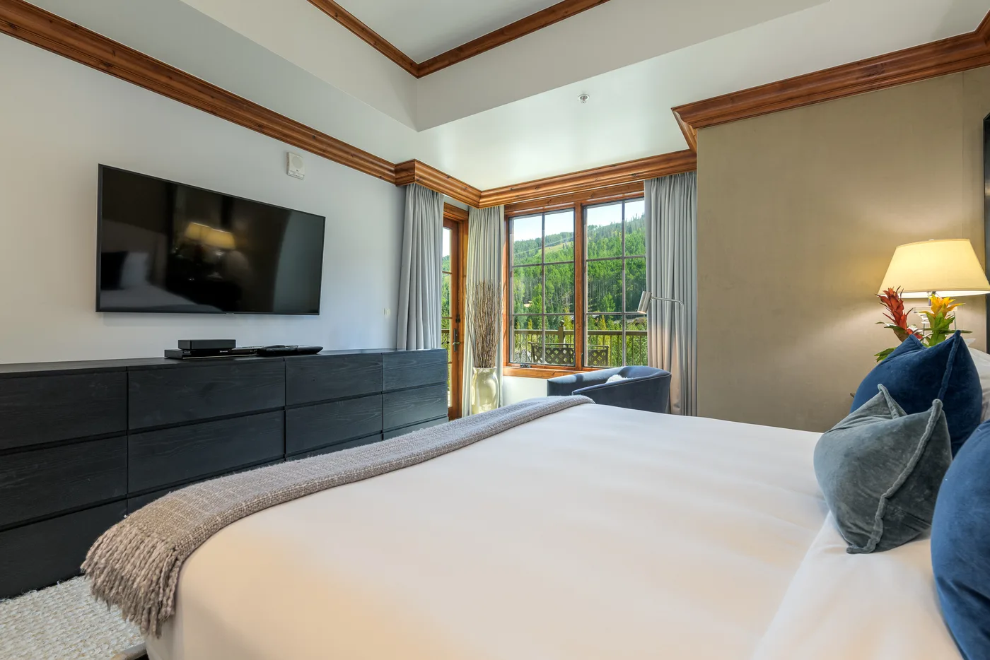 A bedroom with a large bed, a television, and windows overlooking greenery.