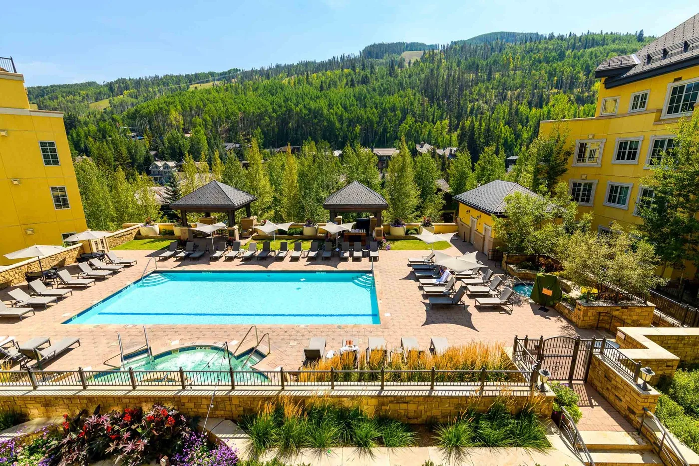 An outdoor pool area with lounge chairs sits among buildings and wooded hills.