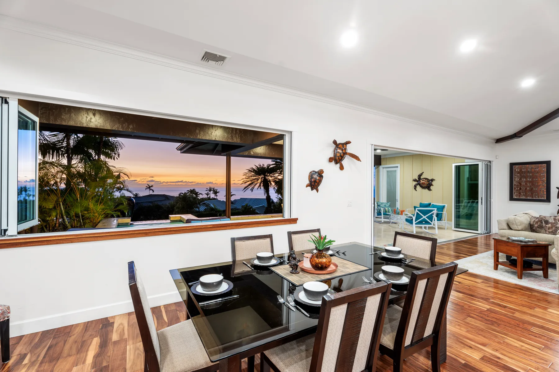 Un comedor con la mesa puesta junto a una ventana amplia con vistas a palmeras y al mar.