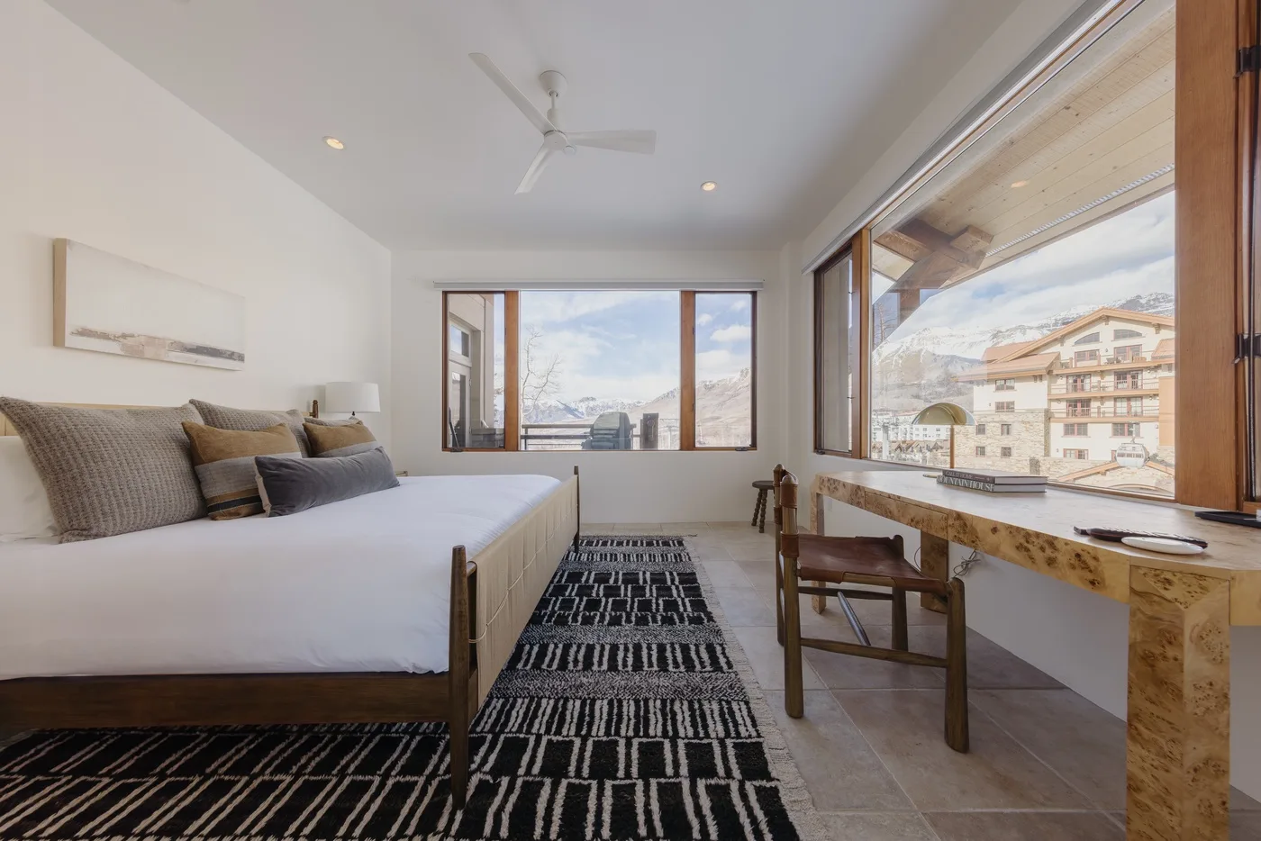 Dormitorio luminoso con cama, alfombra estampada y grandes ventanas con vistas a edificios cercanos.