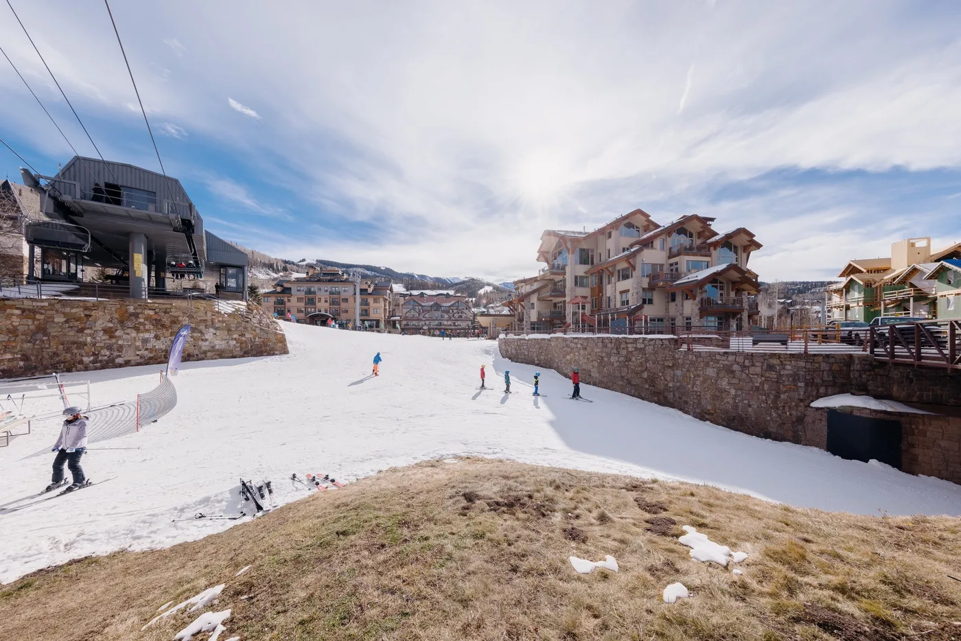 Una zona de esquí nevada con personas esquiando entre edificios y un muro de piedra.