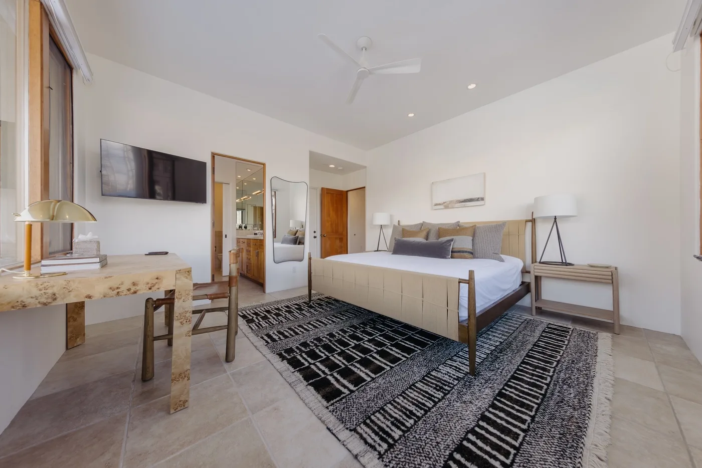 Un dormitorio luminoso con una cama grande, alfombras estampadas y un escritorio junto a la ventana.