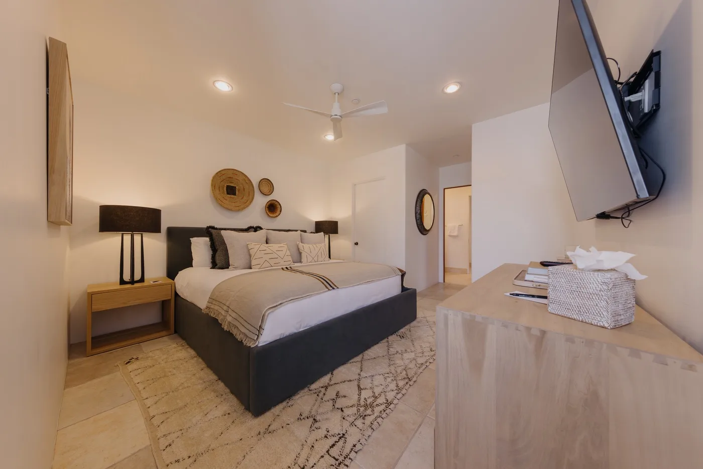 Un dormitorio con una cama tendida y una cómoda de madera bajo un televisor montado en la pared.