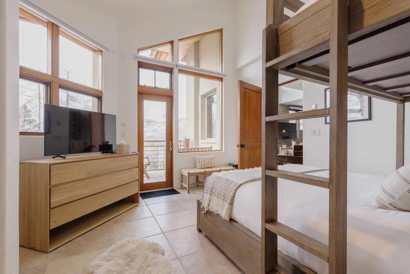 Un dormitorio luminoso con una cama, una cómoda de madera y grandes ventanas.