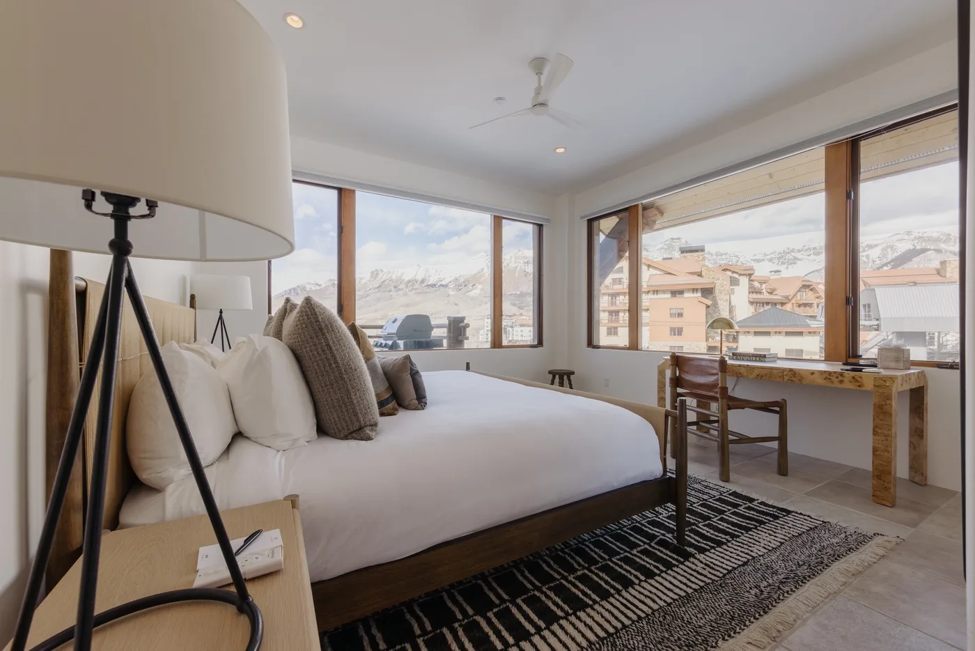 Un dormitorio con una cama tendida junto a grandes ventanales con vistas a edificios cercanos.