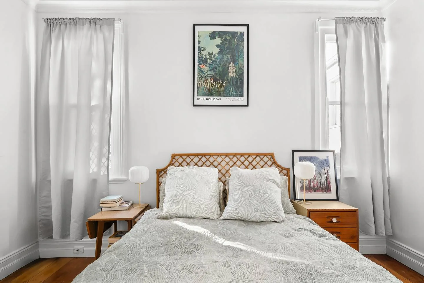 Un dormitorio luminoso con una cama, ventanas con cortinas y cuadros enmarcados.