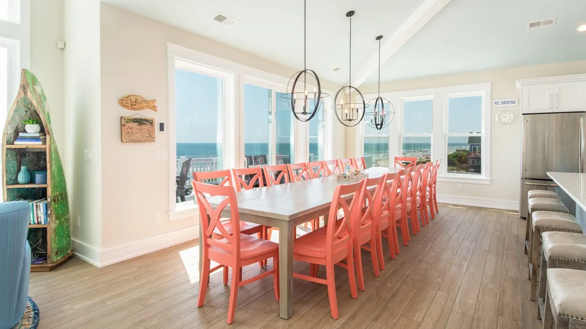 A bright dining room has a long table surrounded by coral-colored seating.