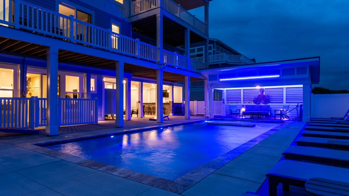 A swimming pool sits beside a multi-level building, illuminated by blue lights at night.