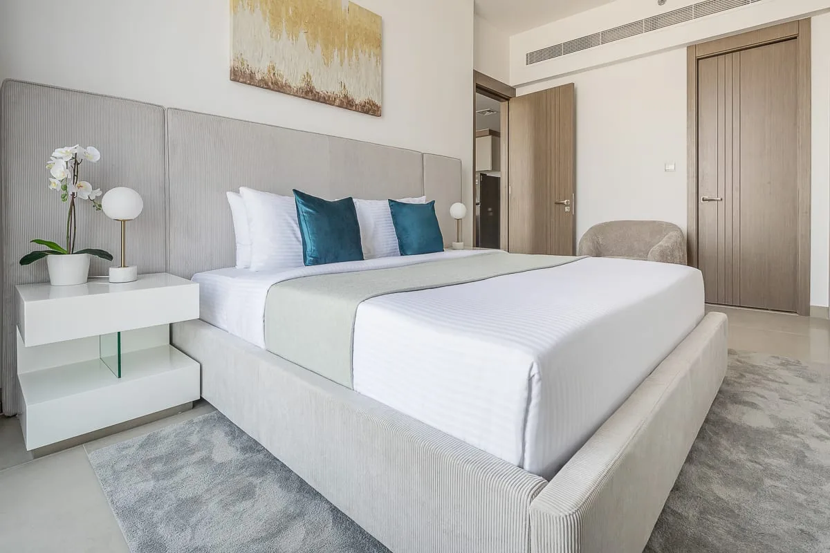 A bright bedroom with a neatly made bed, teal pillows, and a gray rug.