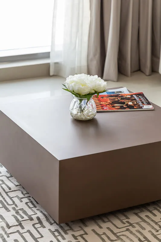 A low coffee table with flowers and magazines sits on a patterned rug by a window.