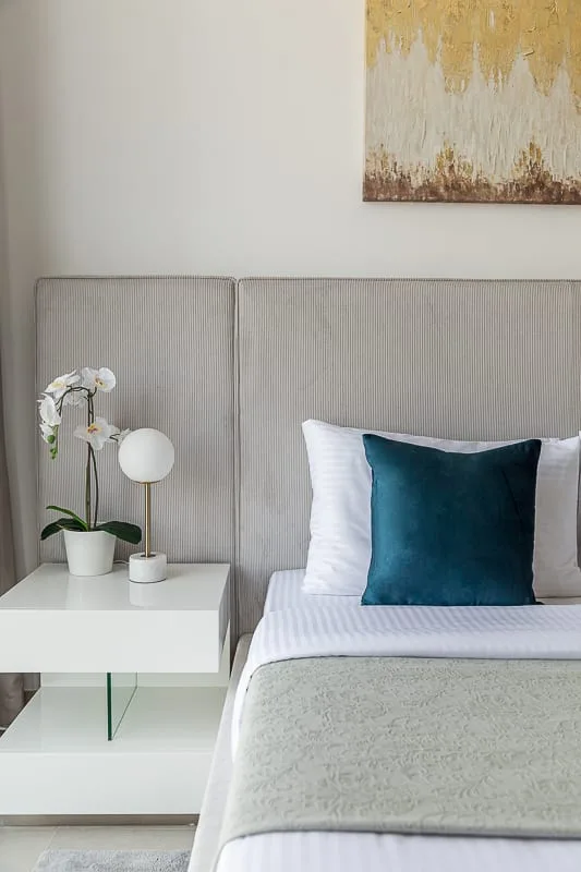 A bedroom with a neatly made bed, teal accent pillow, and bedside table.