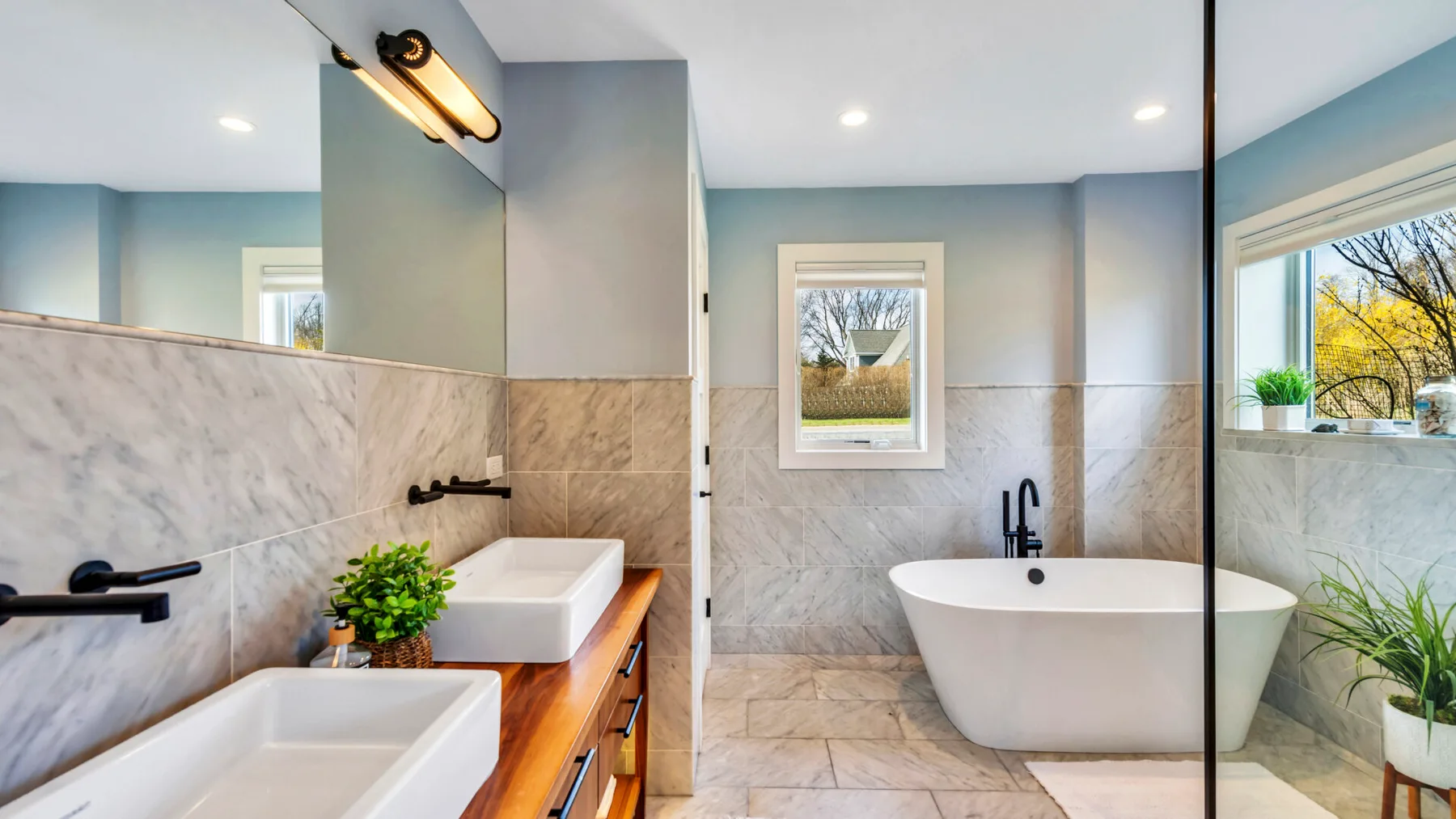 A bright bathroom features a freestanding bathtub, two sinks, and large windows.