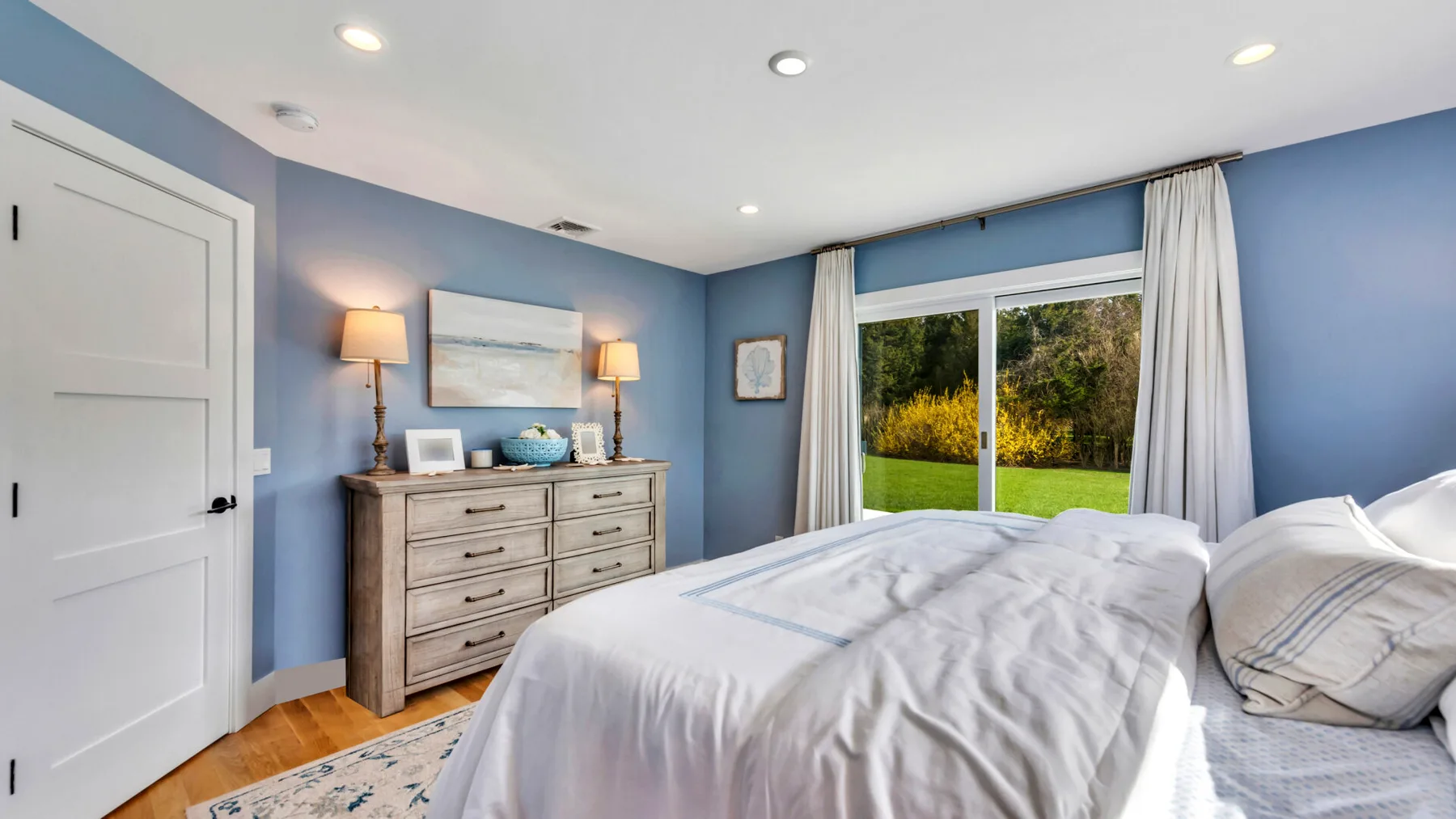 A bedroom with a bed, dresser, and a large window overlooking greenery.