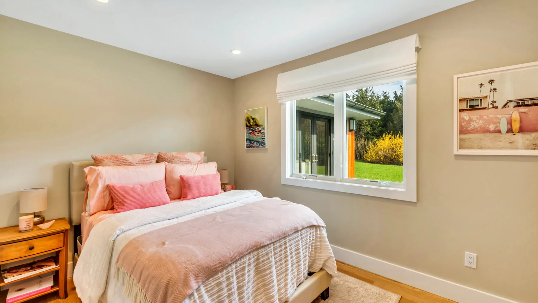 A bedroom with a bed dressed in pale pink bedding beside a bright window.