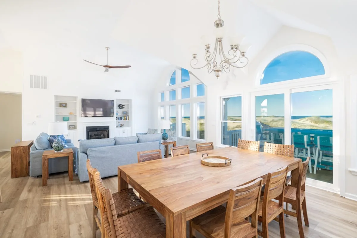 Una luminosa sala de estar y comedor tiene una mesa de madera y grandes ventanales con vistas al agua.