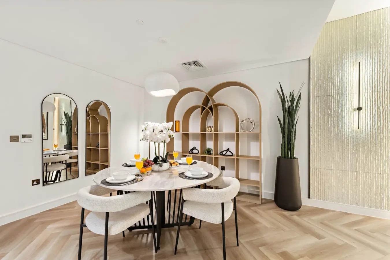 Un comedor luminoso tiene una mesa redonda junto a estanterías y espejos arqueados.