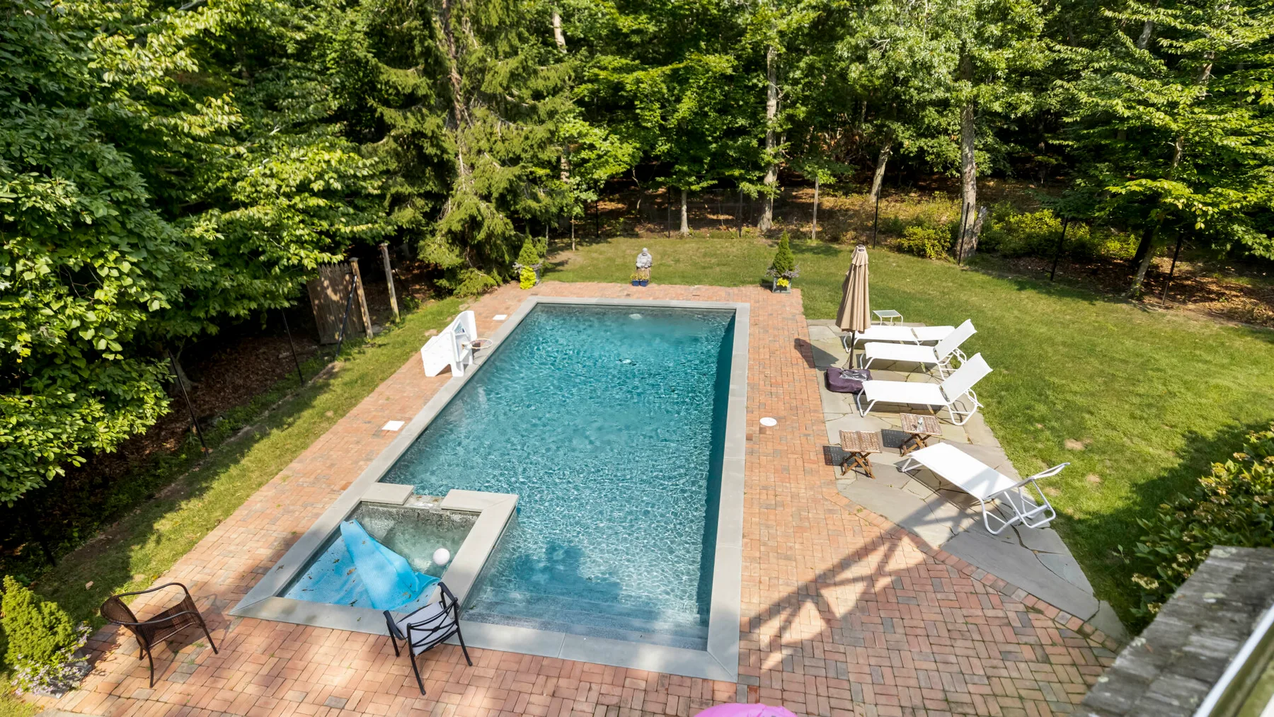 An outdoor swimming pool with a small adjoining spa, surrounded by patio seating and trees.
