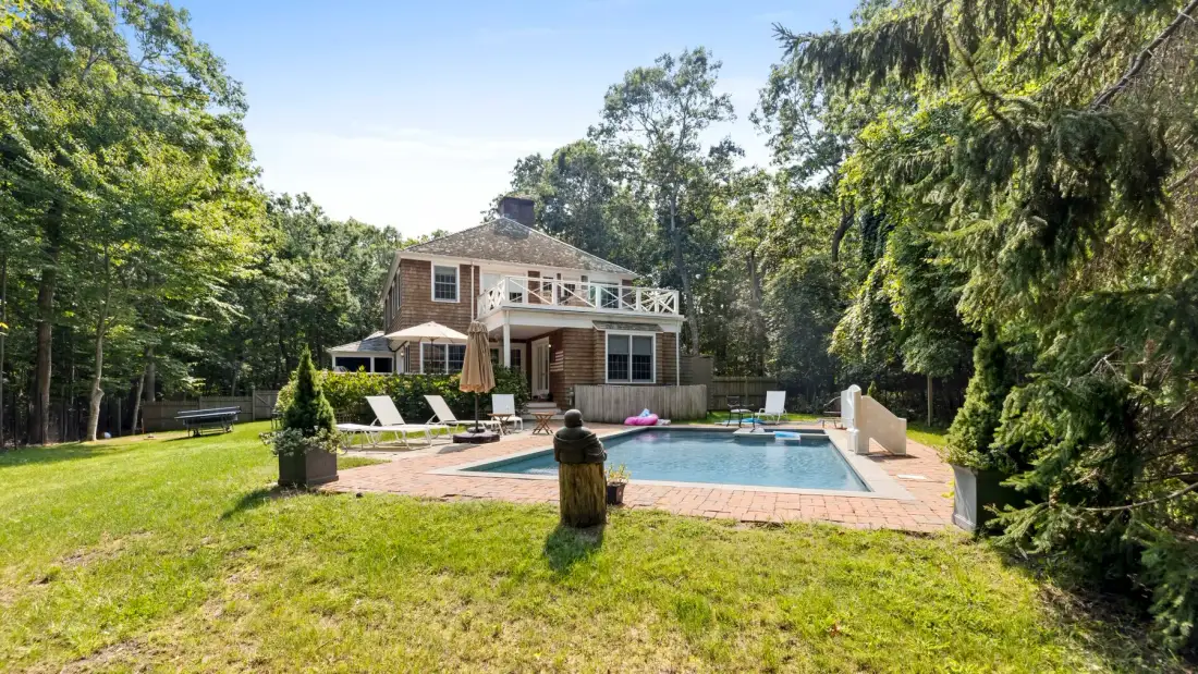 Una casa está junto a una piscina al aire libre, en un jardín arbolado con césped.