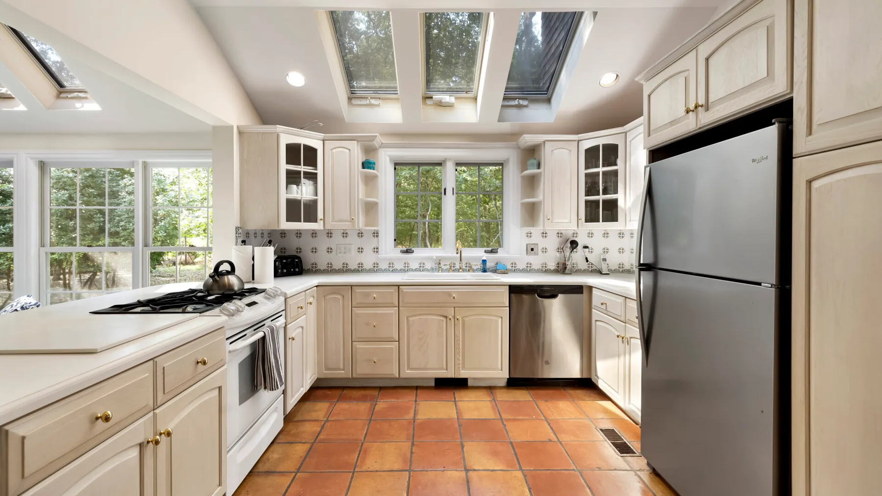 A bright kitchen with pale cabinets, skylights, and a tiled floor.