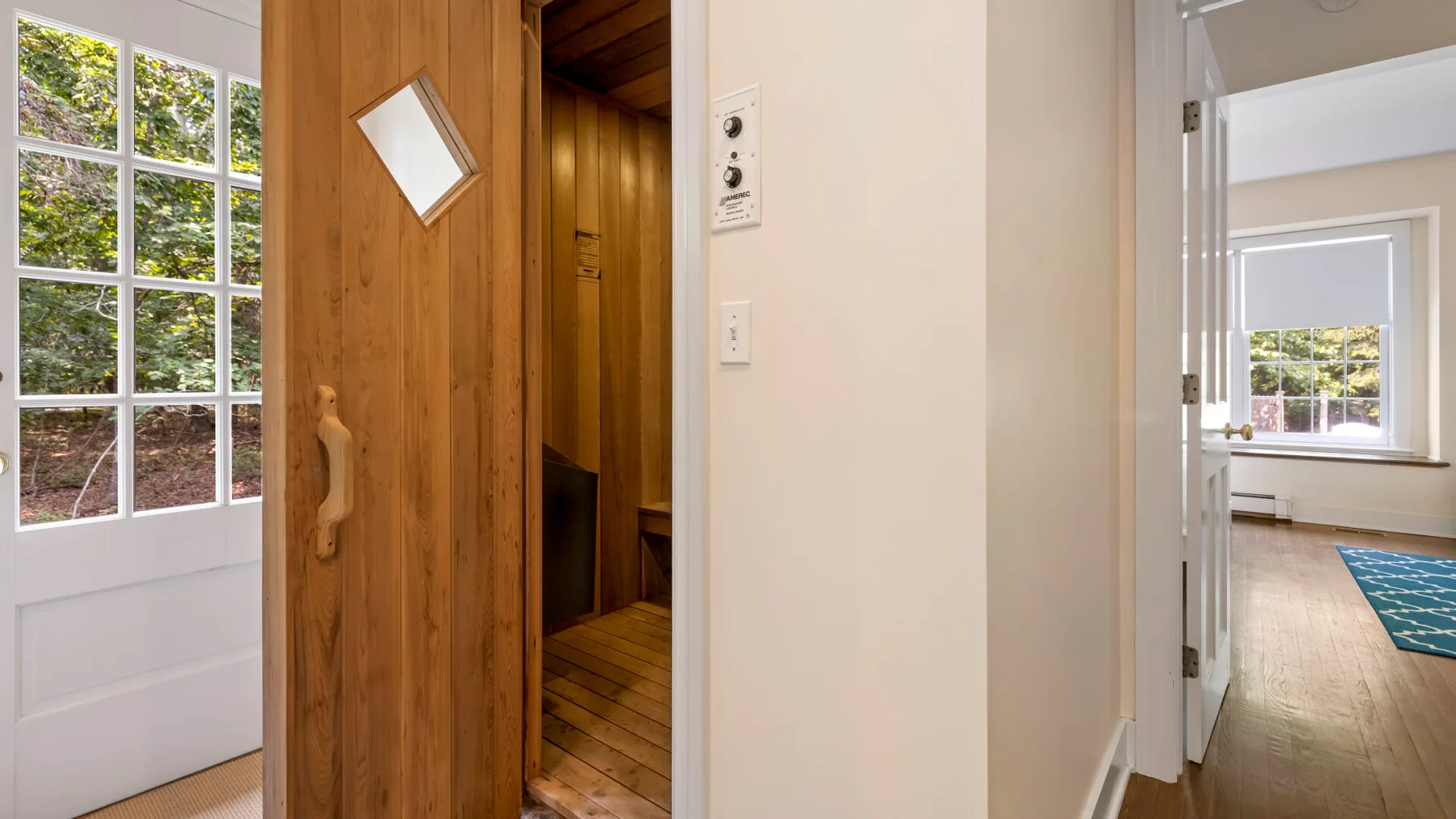 A wooden sauna opens onto a hallway beside a bright room with a window.