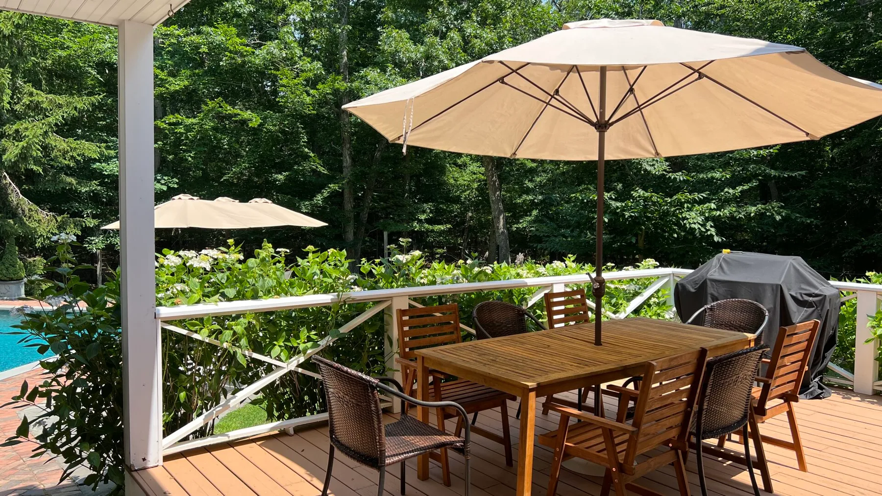 A wooden deck dining area sits beneath a large umbrella beside a swimming pool.