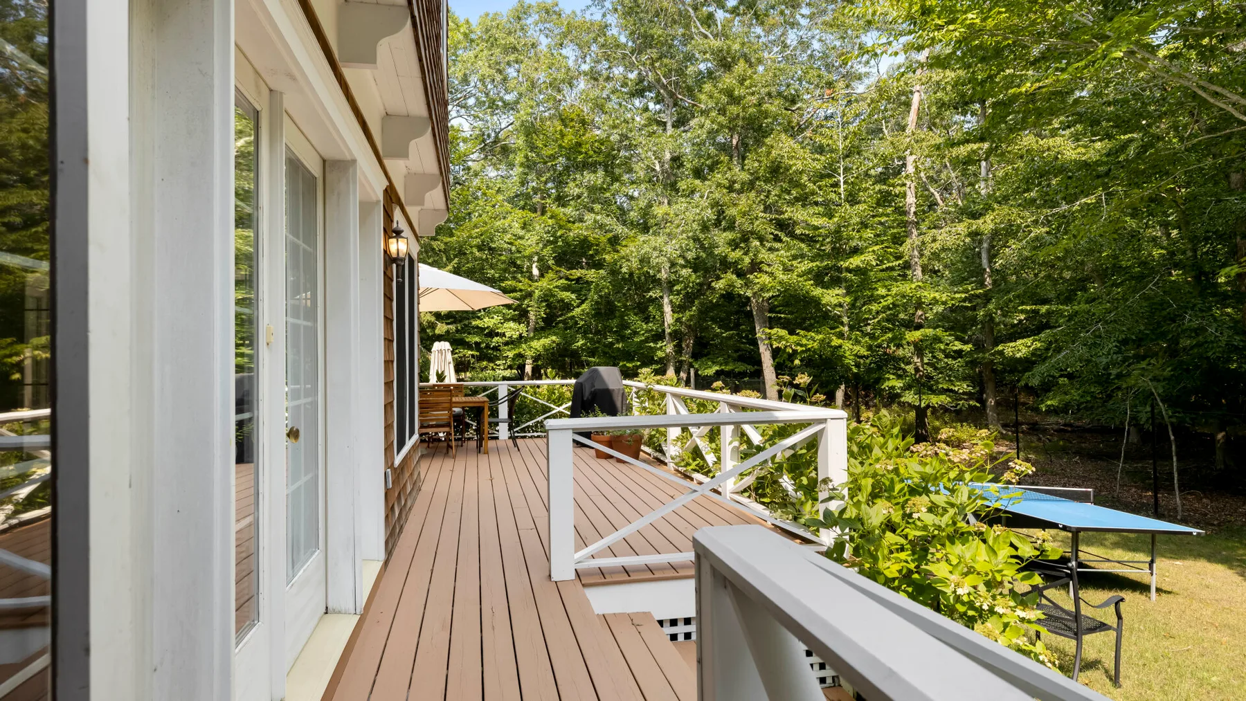 A wooden deck runs alongside a house, with white railings and trees beyond.