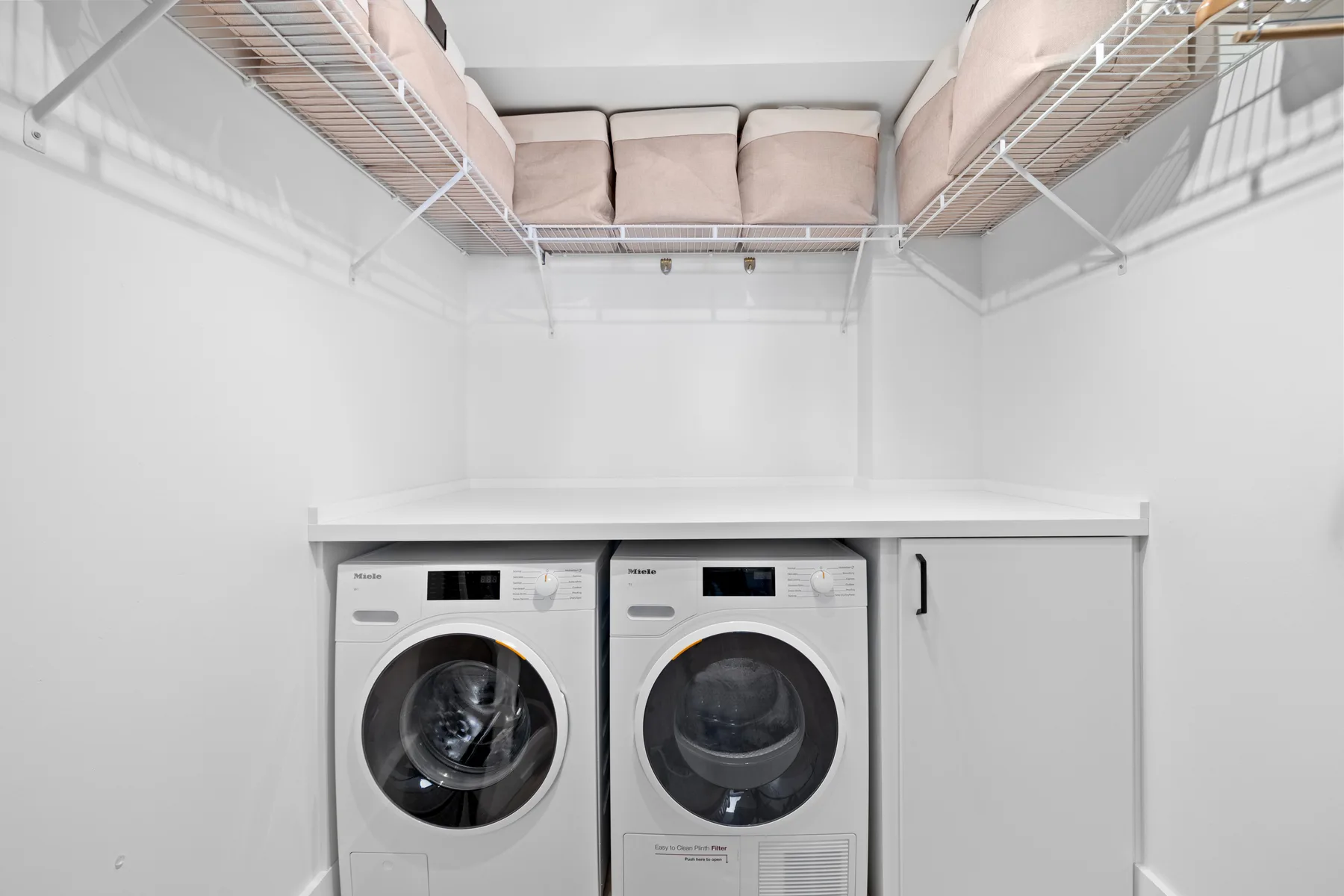 Un cuarto de lavandería con dos máquinas de carga frontal bajo estantes de alambre.