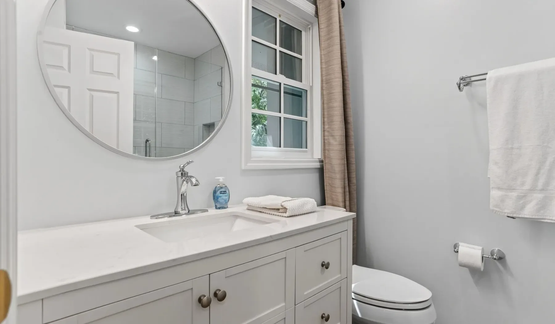 A bright bathroom with a wide vanity, round mirror, and window.