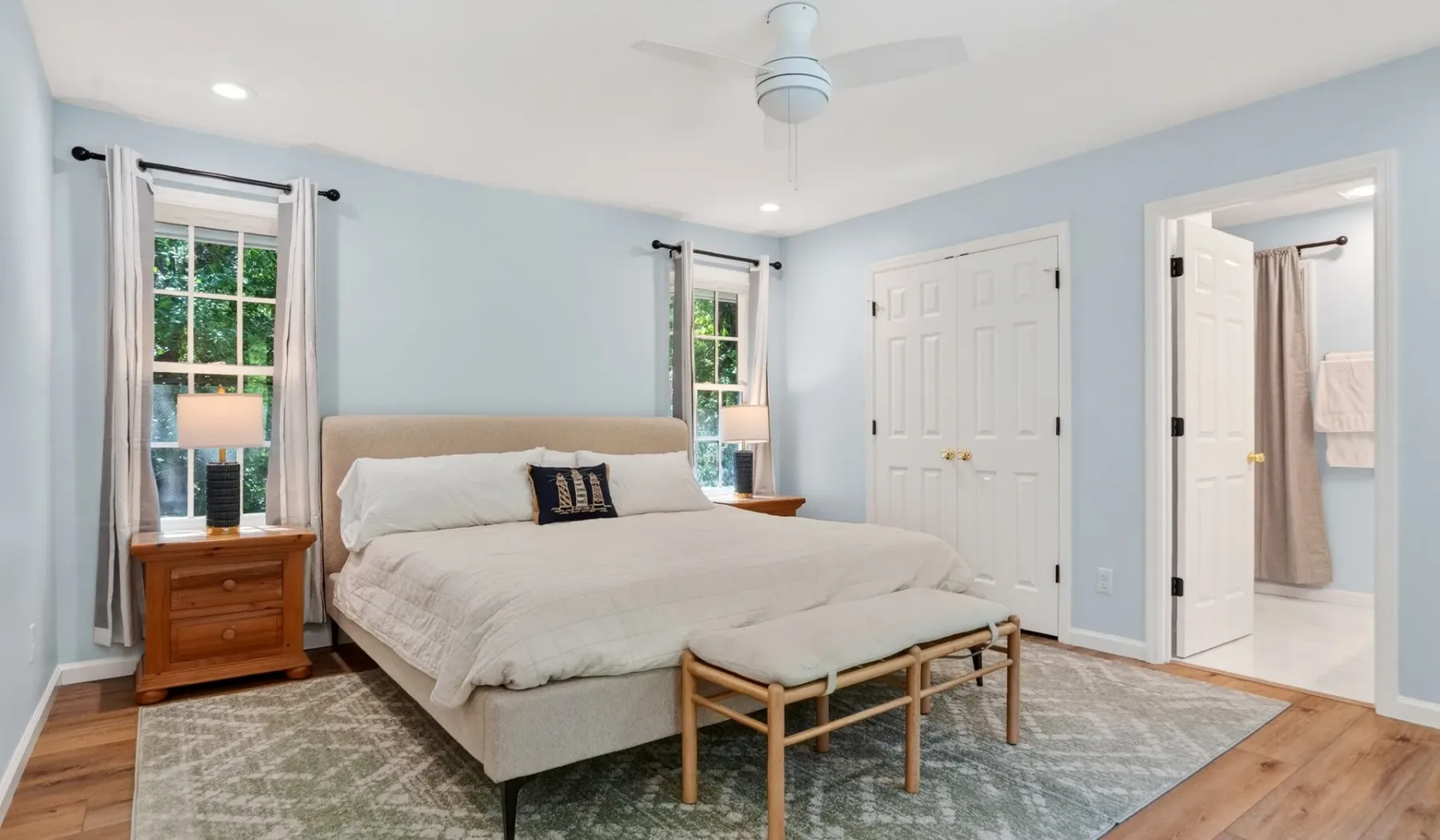 A bedroom with a neatly made bed, bedside table, and windows.