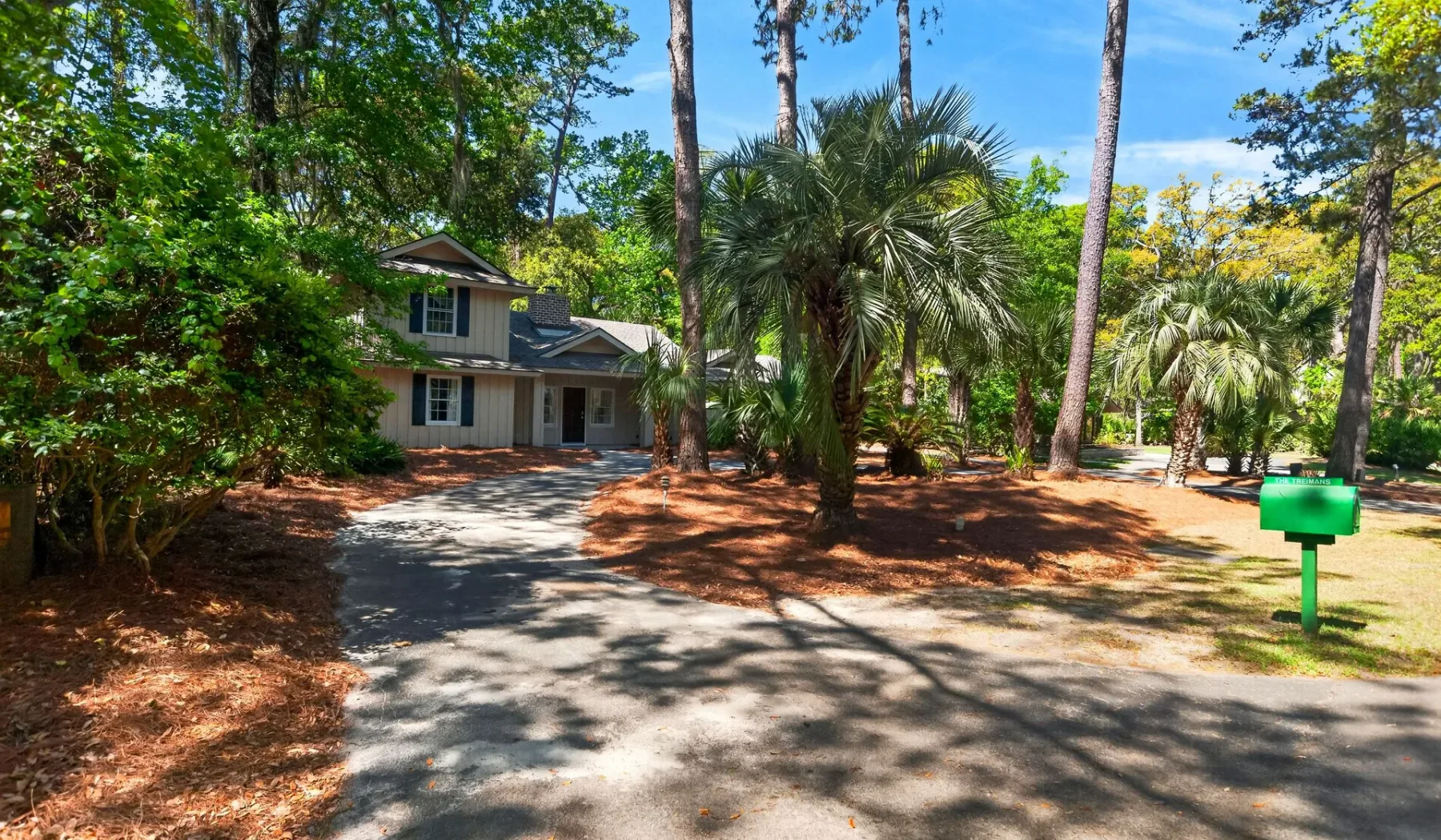 A house stands beside a shaded driveway among mature trees and palms.