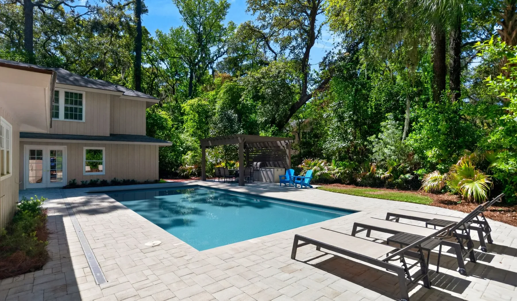 An outdoor swimming pool beside a paved patio, with lounge seating and trees.