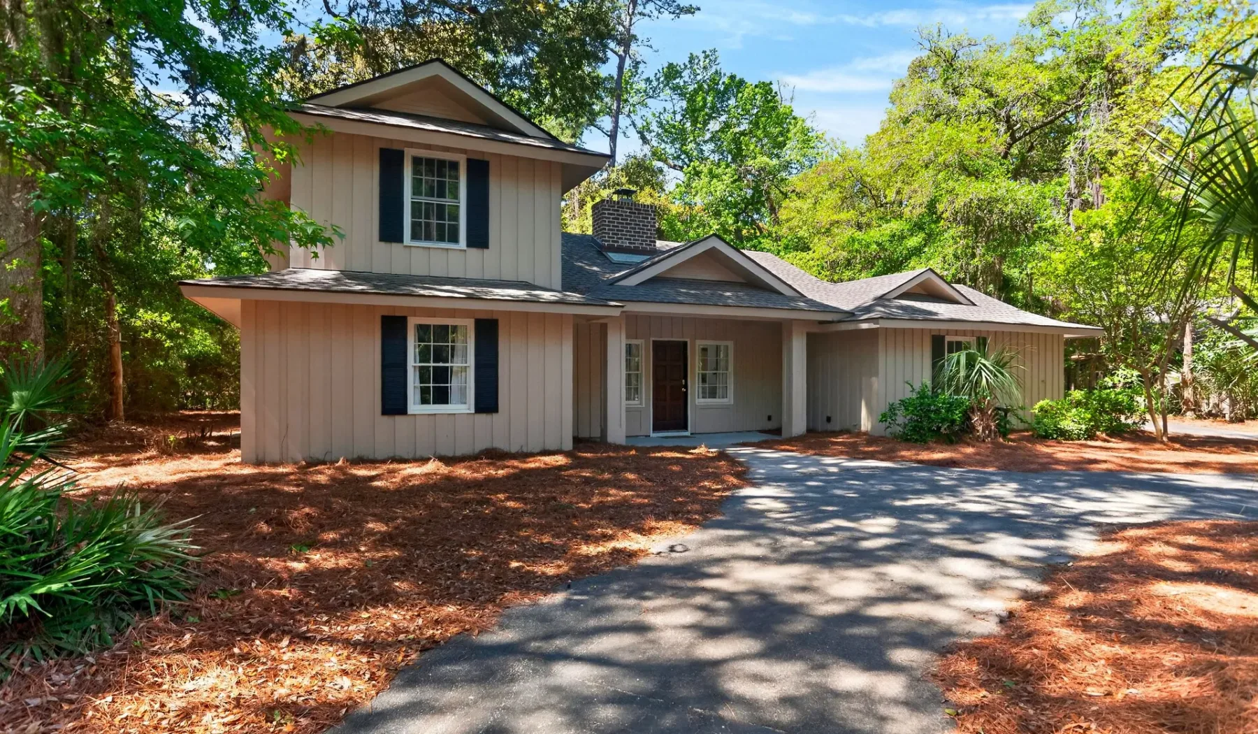 A light-colored house with a paved driveway surrounded by trees.