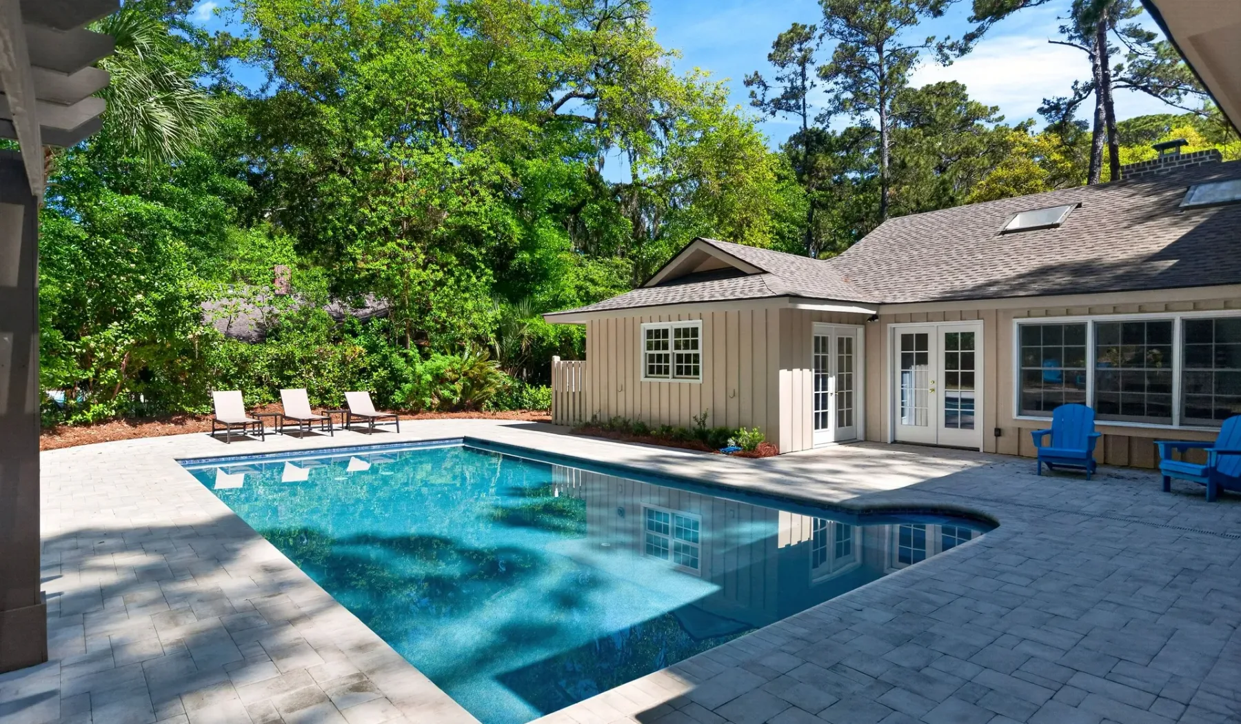 An outdoor swimming pool beside a house, surrounded by trees and patio seating.