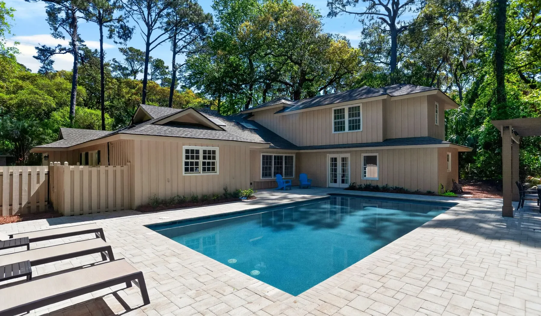 A house stands beside a swimming pool surrounded by paved patio space and trees.