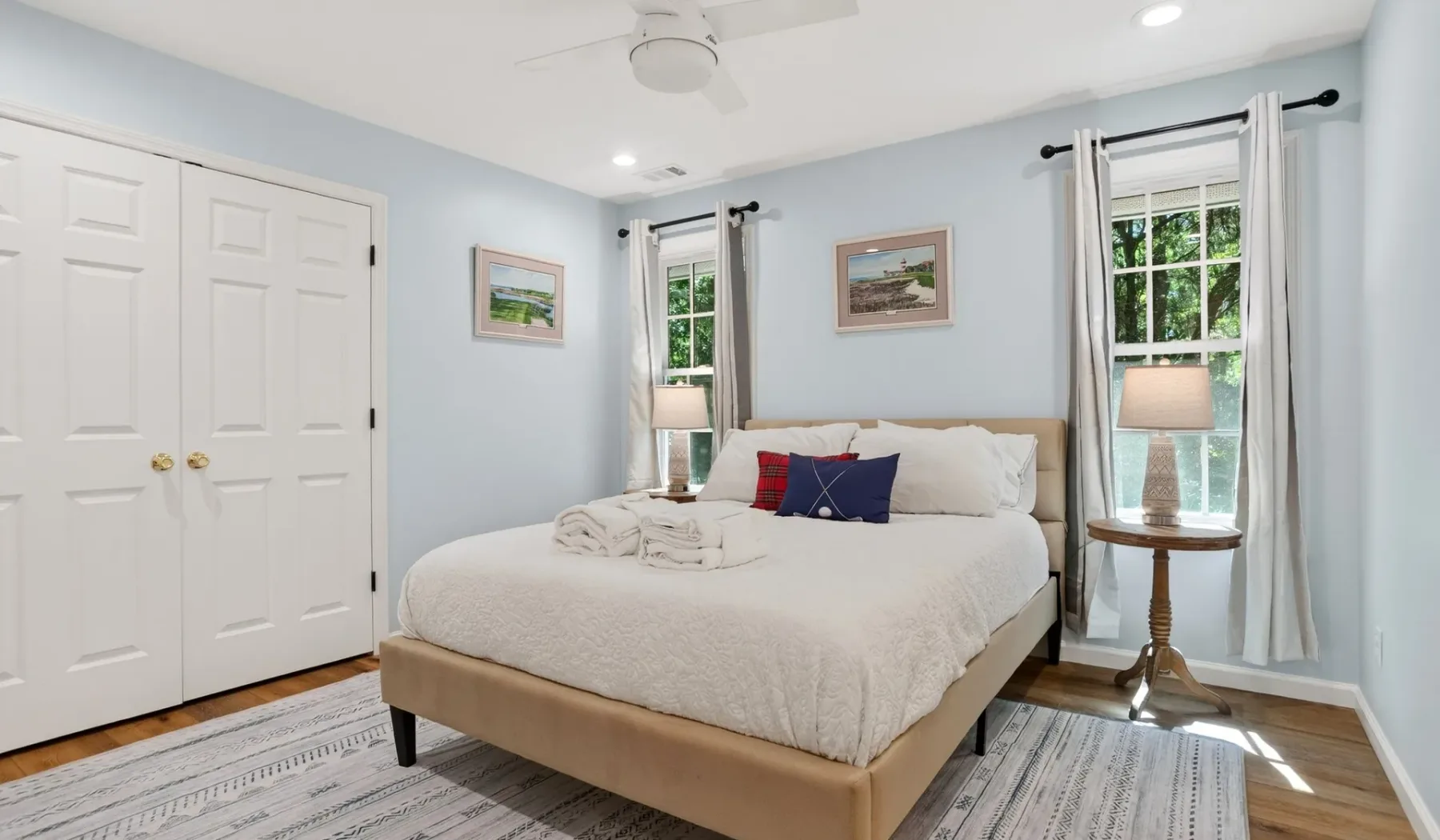 A bright bedroom with a neatly made bed, framed pictures, and curtained windows.