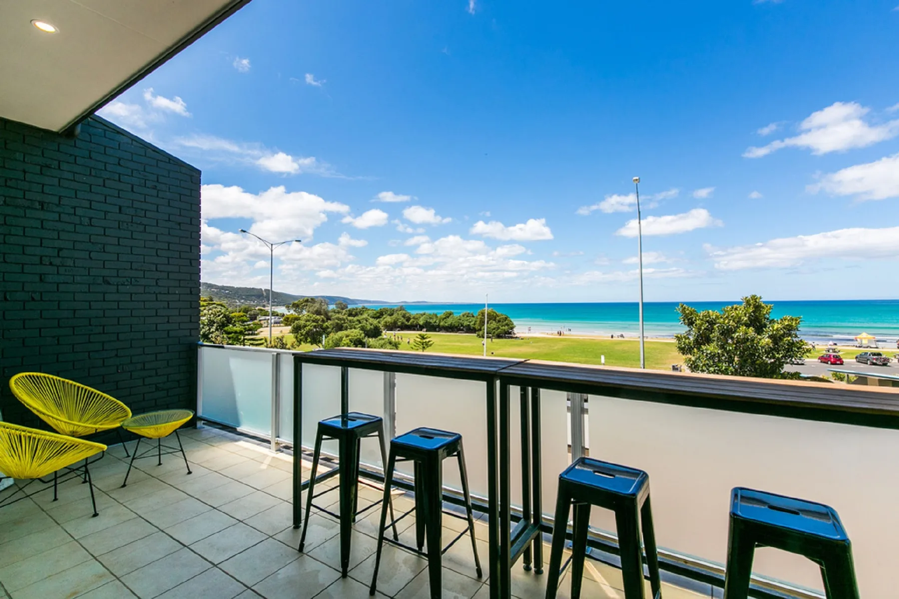 A balcony seating area overlooks a green park and the ocean.
