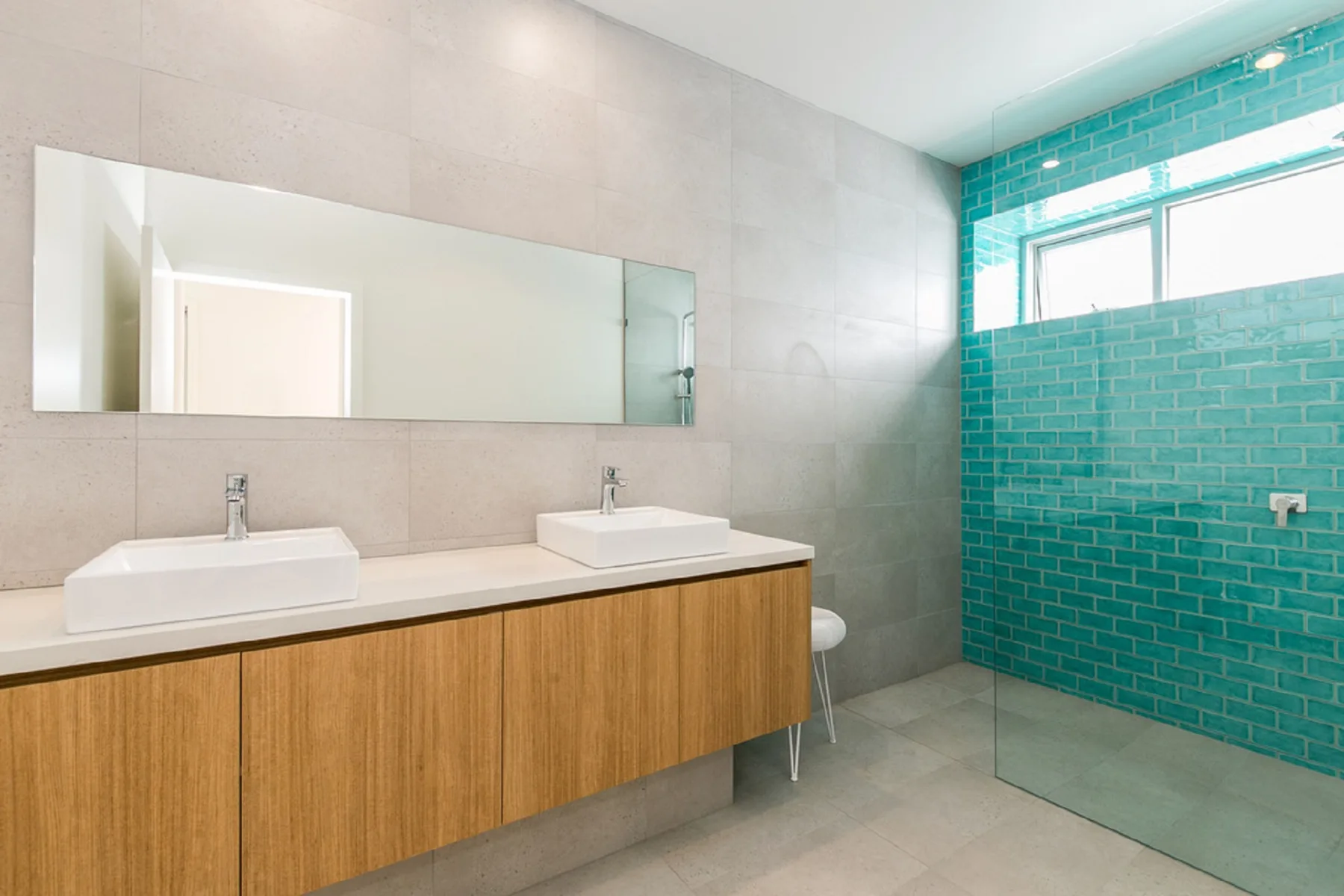 A modern bathroom with a long vanity, two sinks, and a turquoise tiled shower area.