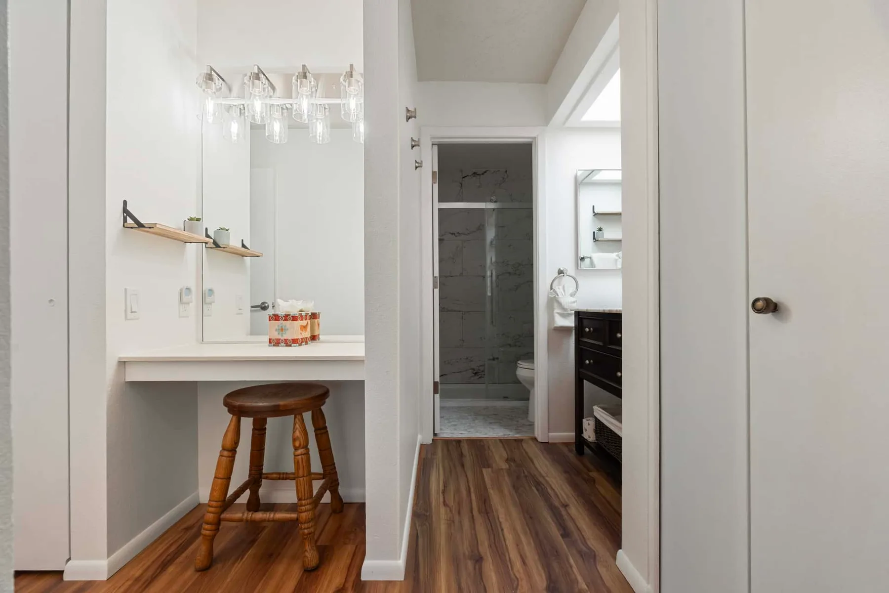 A bright bathroom vanity area opens onto a shower and a wood-floored hallway.