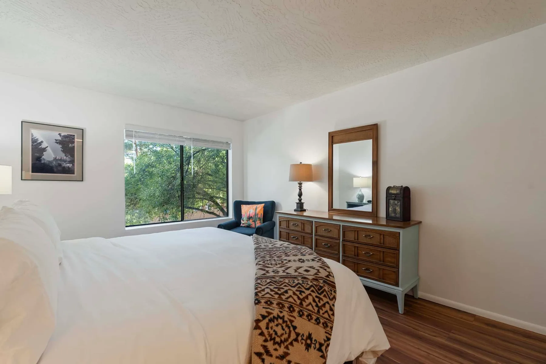 A bedroom with a large bed, a dresser, and a window overlooking trees.