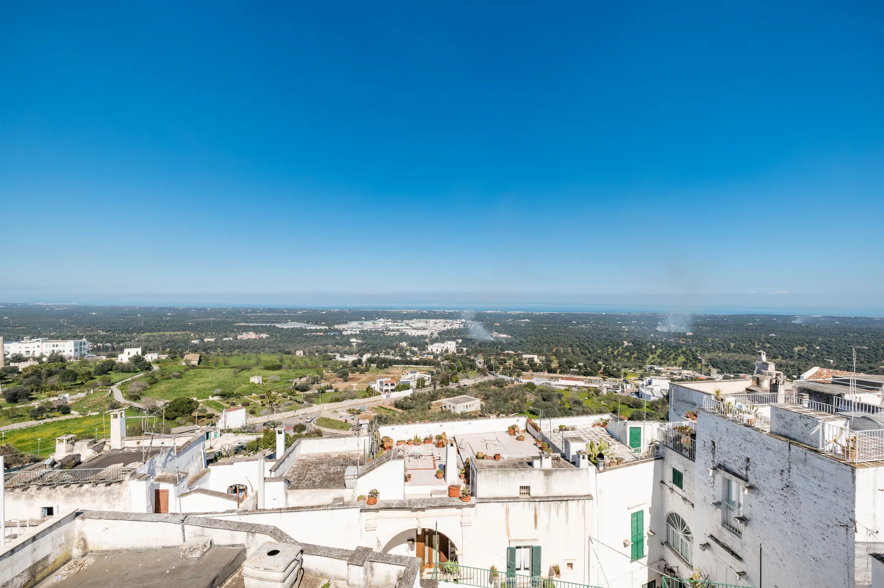 Una vista desde una azotea muestra edificios blancos y un paisaje verde bajo un cielo azul despejado.
