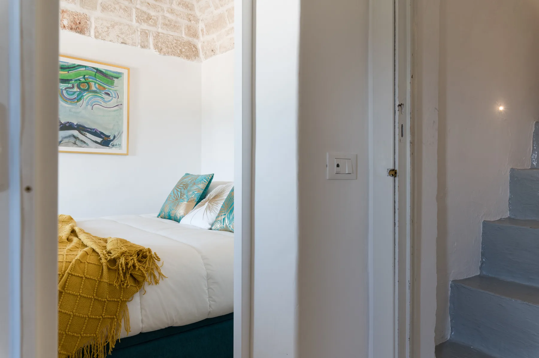 Un dormitorio con una cama blanca, cojines coloridos y una obra enmarcada.