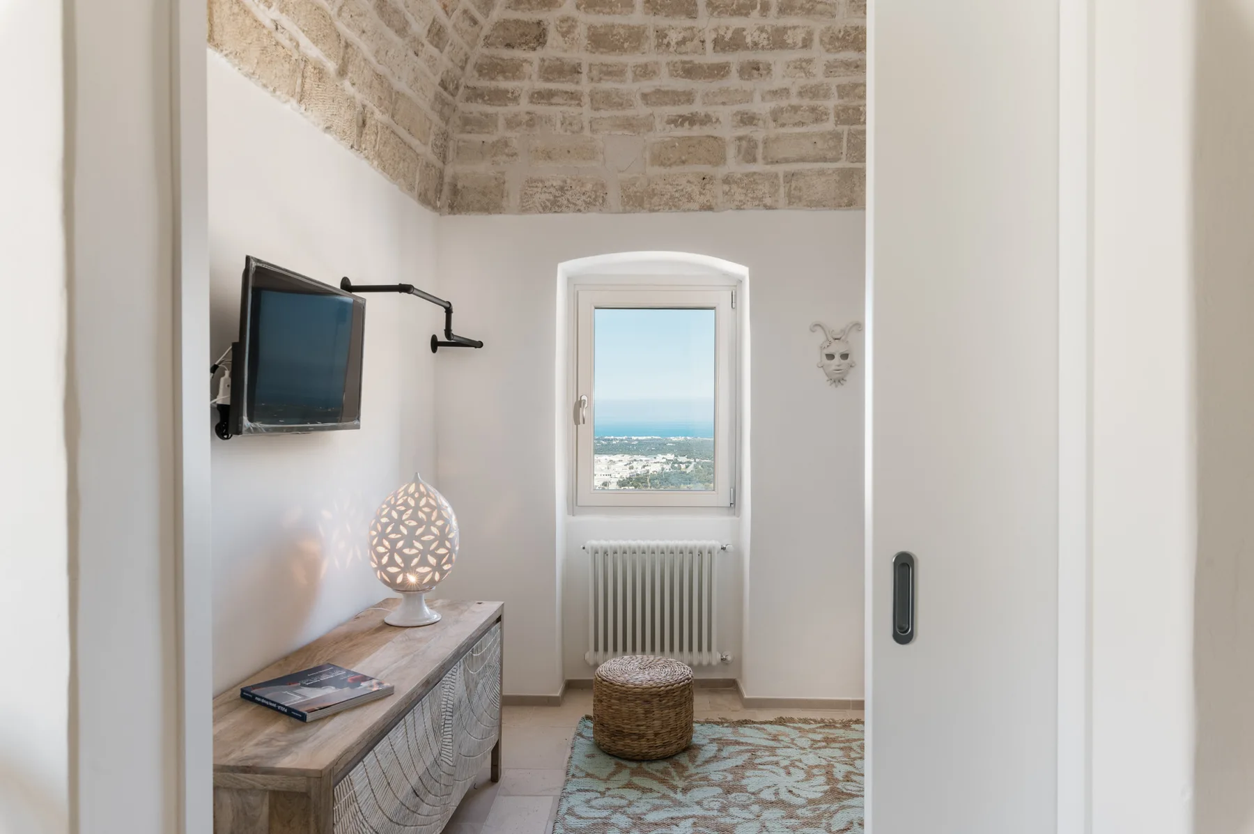 Una habitación luminosa con un televisor en la pared, una ventana y una pequeña zona para sentarse.