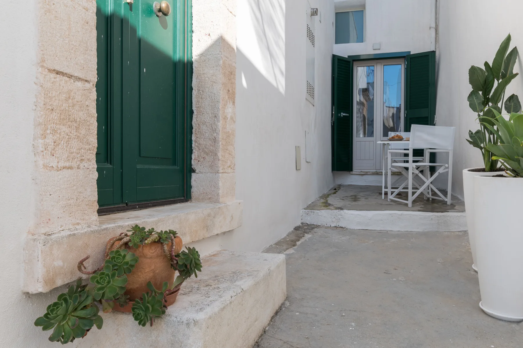 Un patio de paredes blancas con puertas verdes, asientos al aire libre y plantas en macetas.