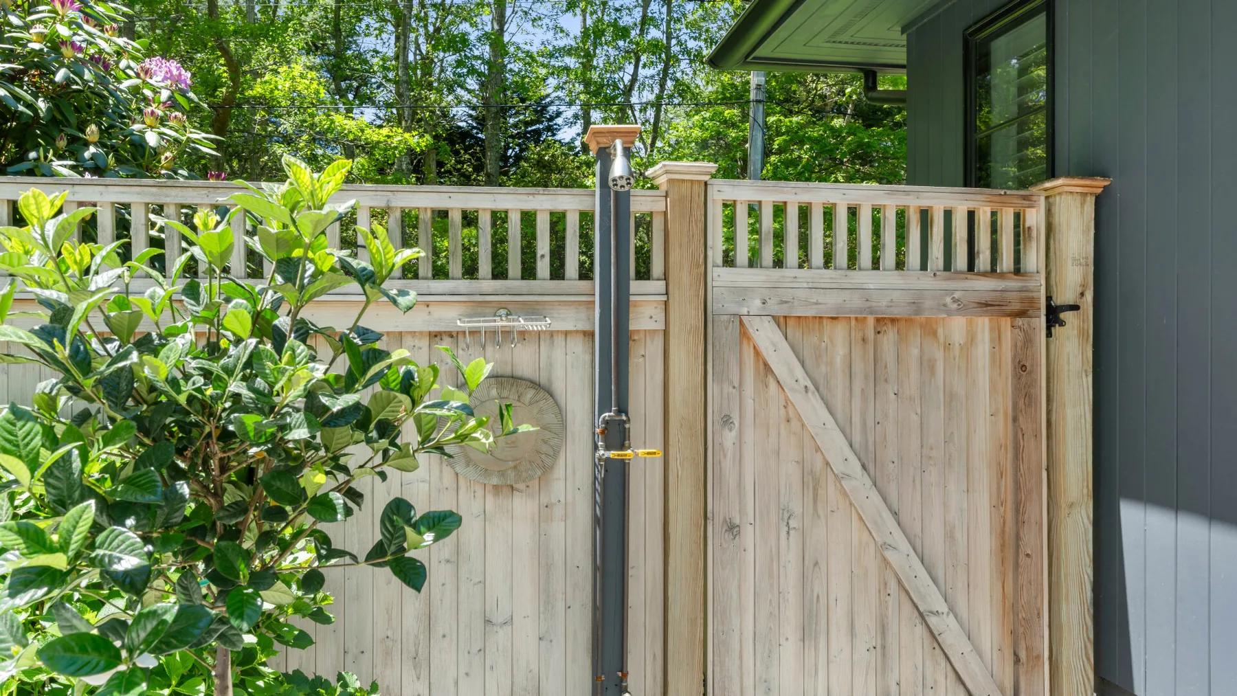 An outdoor shower stands beside a wooden privacy fence and leafy plants.