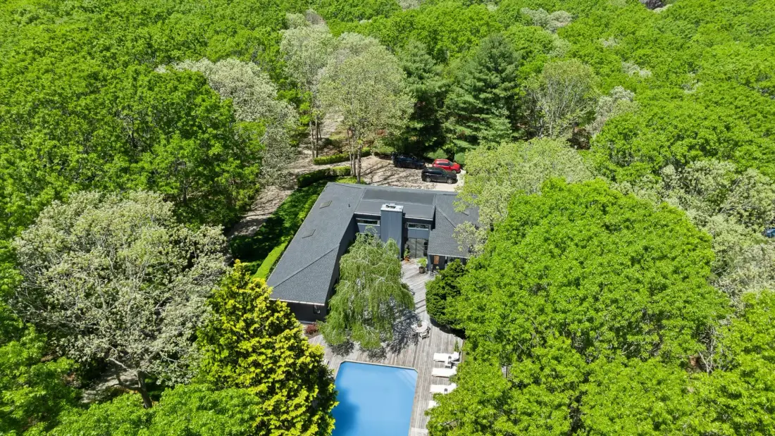 An aerial view of a house surrounded by dense green trees, with a swimming pool.