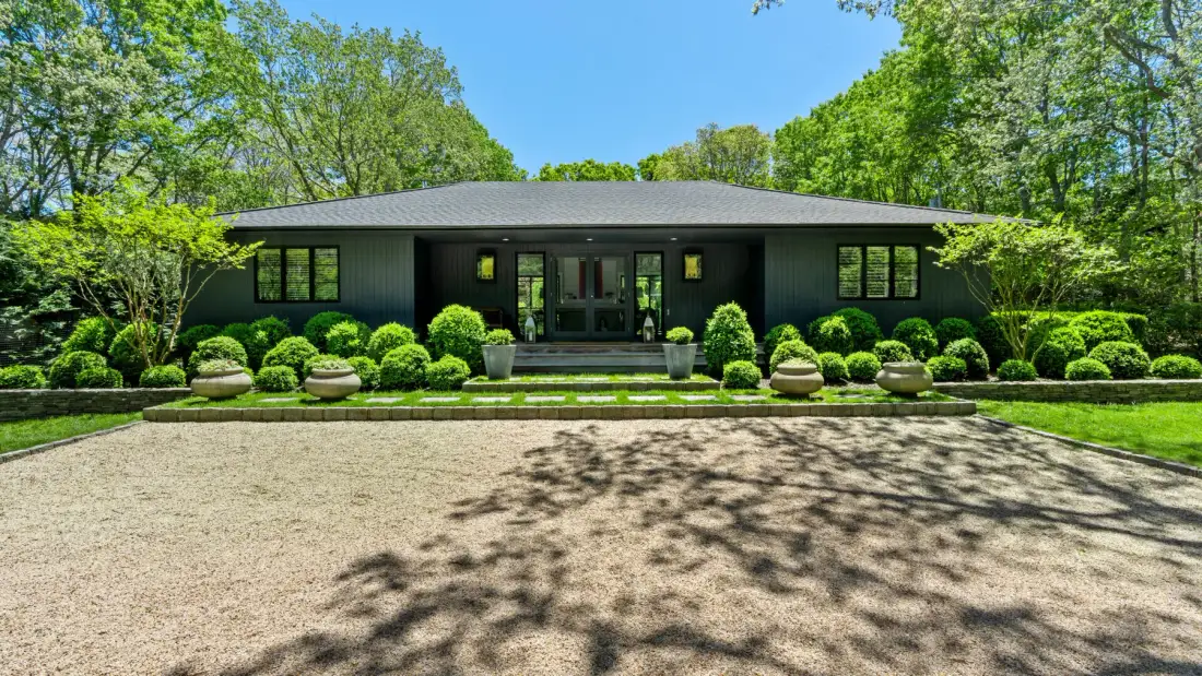 A modern single-story house sits behind a broad gravel driveway and manicured shrubs.