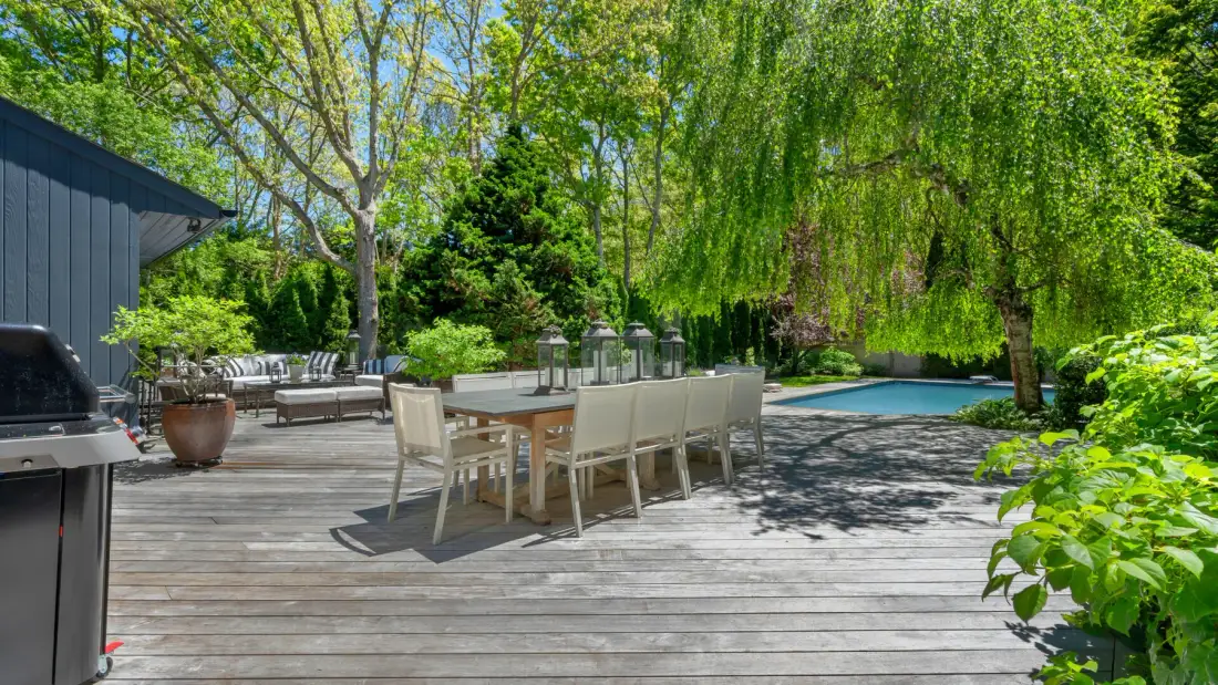 An outdoor dining area on a wooden deck beside a swimming pool and leafy trees.