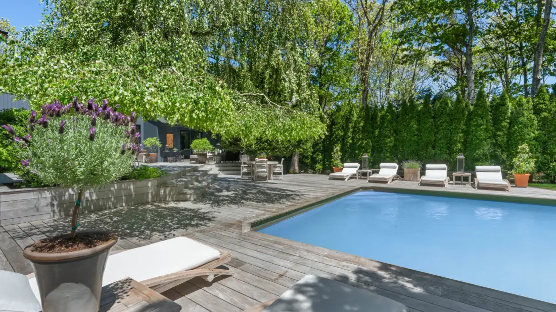 An outdoor swimming pool sits beside a wooden deck with lounge seating and trees.