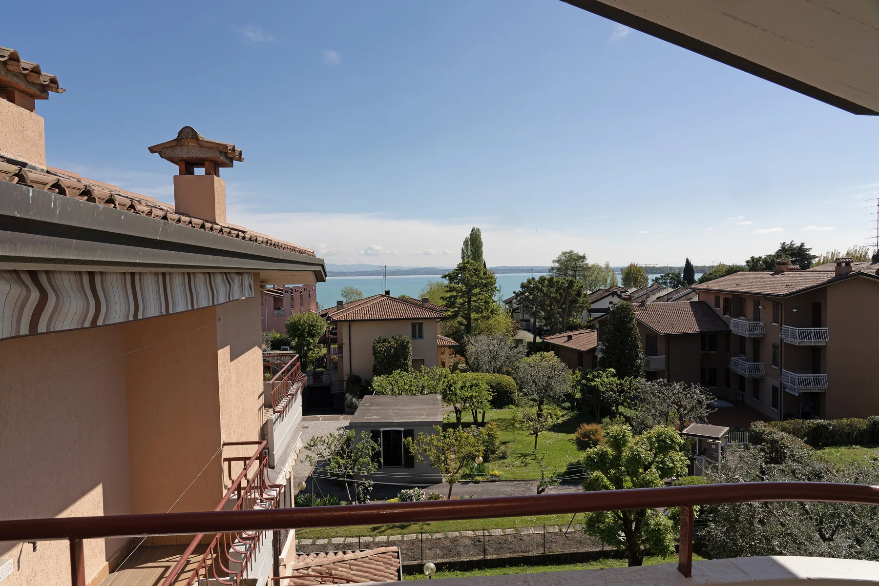 A balcony overlooks a garden, nearby buildings, and a distant lake.